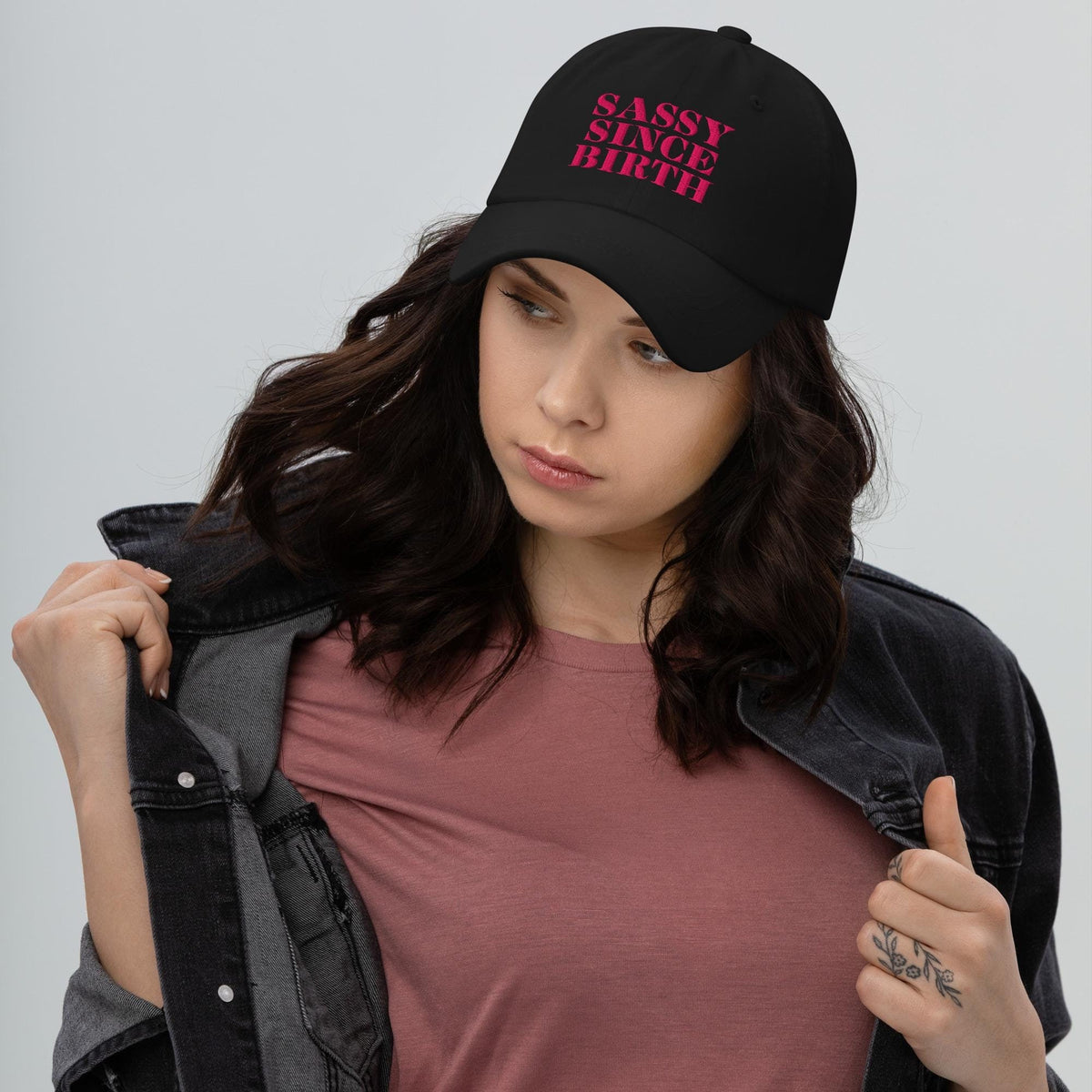 Black low-profile dad cap worn by a woman, embroidered in pink with the words Sassy Since Birth.