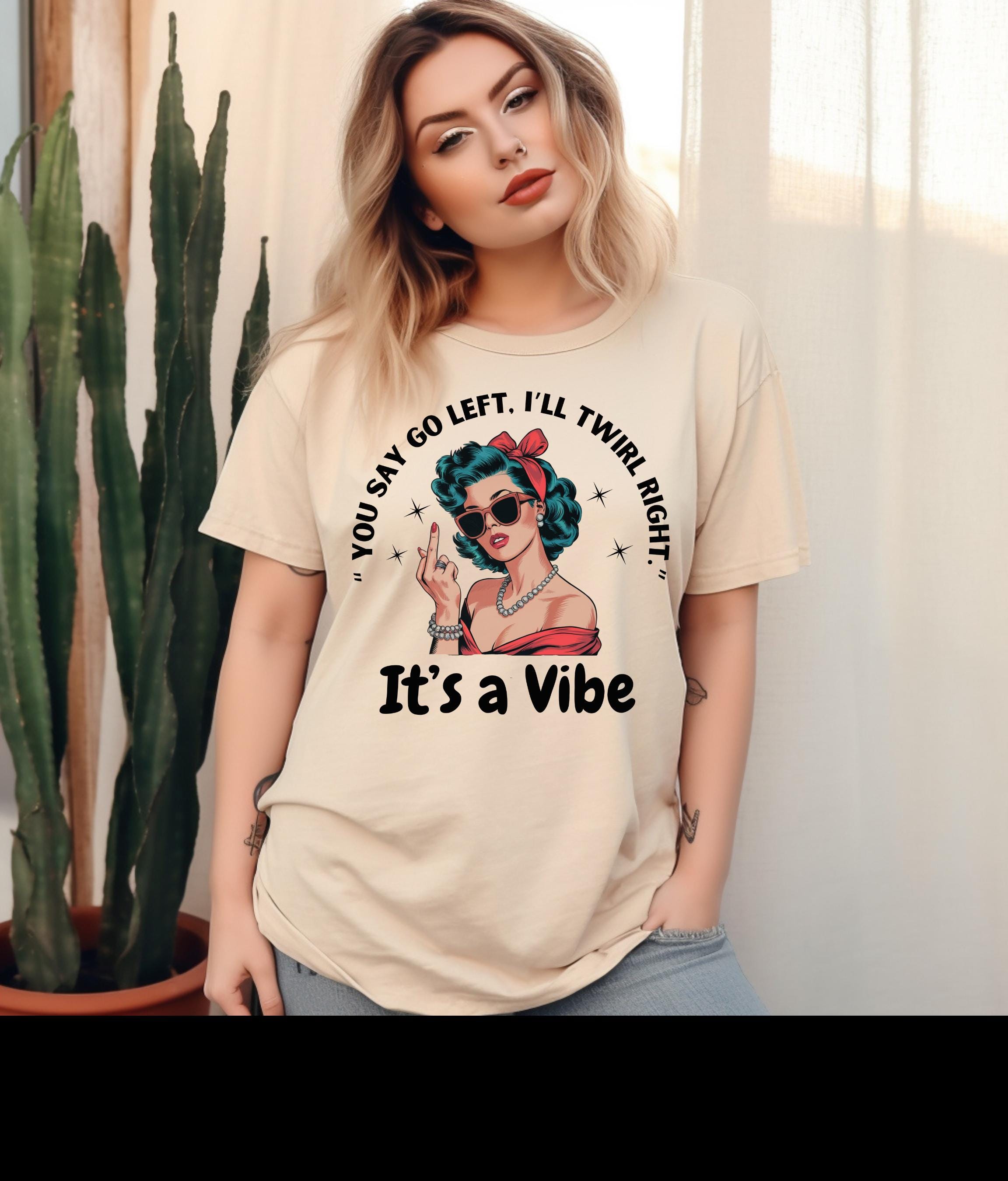 May include: Beige t-shirt with a retro illustration of a woman in sunglasses and a red bow, giving the middle finger. The text on the shirt reads "You say go left, I'll twirl right." and "It's a Vibe."