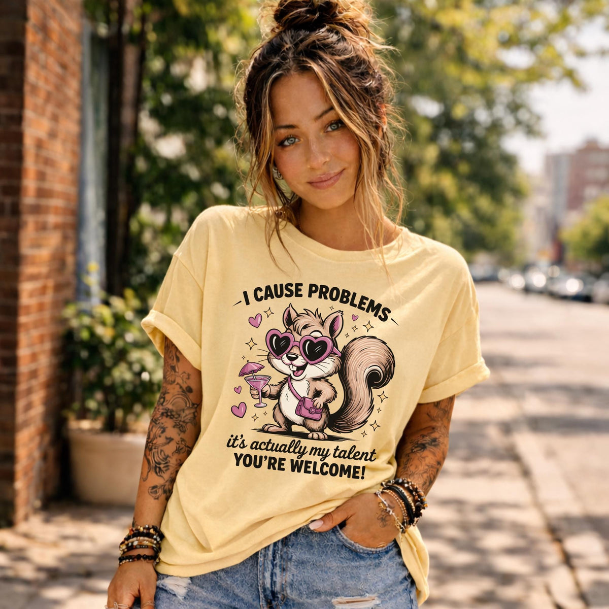 Woman wearing a light yellow graphic t-shirt featuring a cute squirrel with heart sunglasses and drink, with the text “I Cause Problems It’s Actually My Talent You’re Welcome,” styled in a sunny outdoor setting.