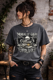 Pepper graphic t-shirt flat lay featuring “Morally Gray Book Club” design with a skull resting on stacked books, gothic ornamental details, and a glowing eye element, styled with books, coffee, and white roses for a dark academia aesthetic.