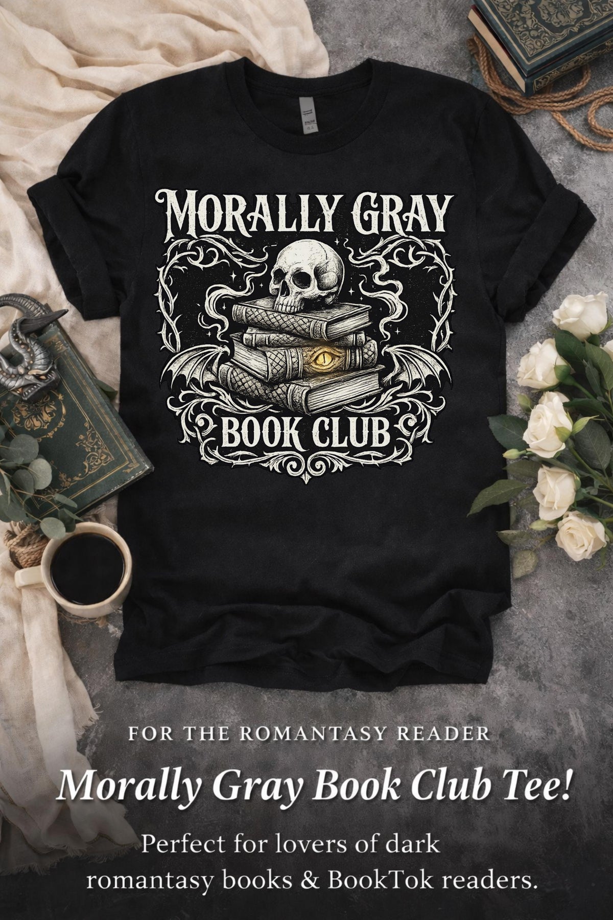 Black graphic t-shirt flat lay featuring “Morally Gray Book Club” design with a skull resting on stacked books, gothic ornamental details, and a glowing eye element, styled with books, coffee, and white roses for a dark academia aesthetic.