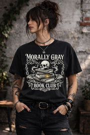 Black graphic t-shirt flat lay featuring “Morally Gray Book Club” design with a skull resting on stacked books, gothic ornamental details, and a glowing eye element, styled with books, coffee, and white roses for a dark academia aesthetic.