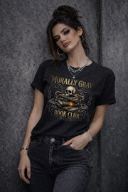 Woman wearing a black “Morally Gray Book Club” graphic t-shirt featuring a skull on stacked books with a glowing eye, styled in a dark academia aesthetic against a moody textured background.