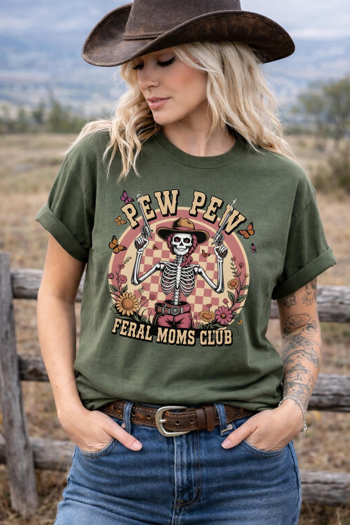 Western graphic t-shirt with skeleton cowgirl holding guns, “Pew Pew Feral Moms Club” text, and vintage floral design, worn in a country-style outdoor setting.
