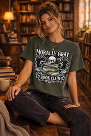 Graphic t-shirt flat lay featuring “Morally Gray Book Club” design with a skull resting on stacked books, gothic ornamental details, and a glowing eye element, styled with books, coffee, and white roses for a dark academia aesthetic.