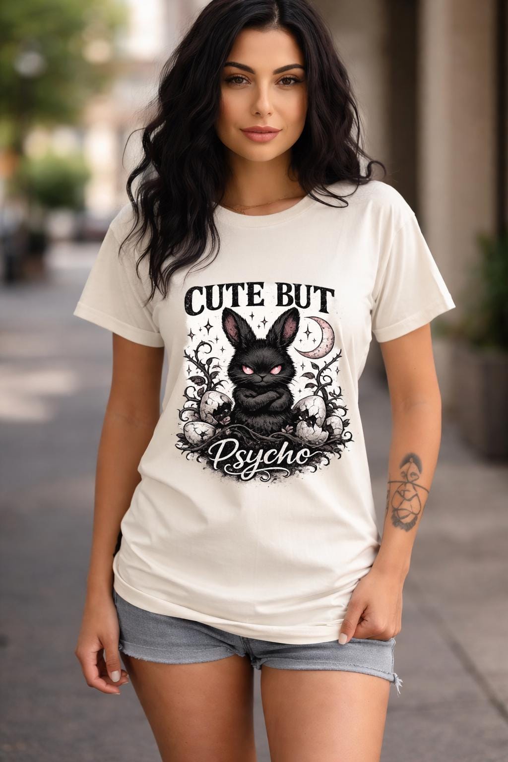 Graphic t-shirt featuring gothic bunny with glowing eyes and “Cute But Psycho” text, worn by model on beach with casual summer outfit