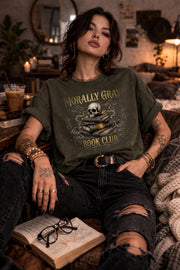 Woman wearing a “Morally Gray Book Club” graphic t-shirt featuring a skull on stacked books with a glowing eye, styled in a dark academia aesthetic against a moody textured background.