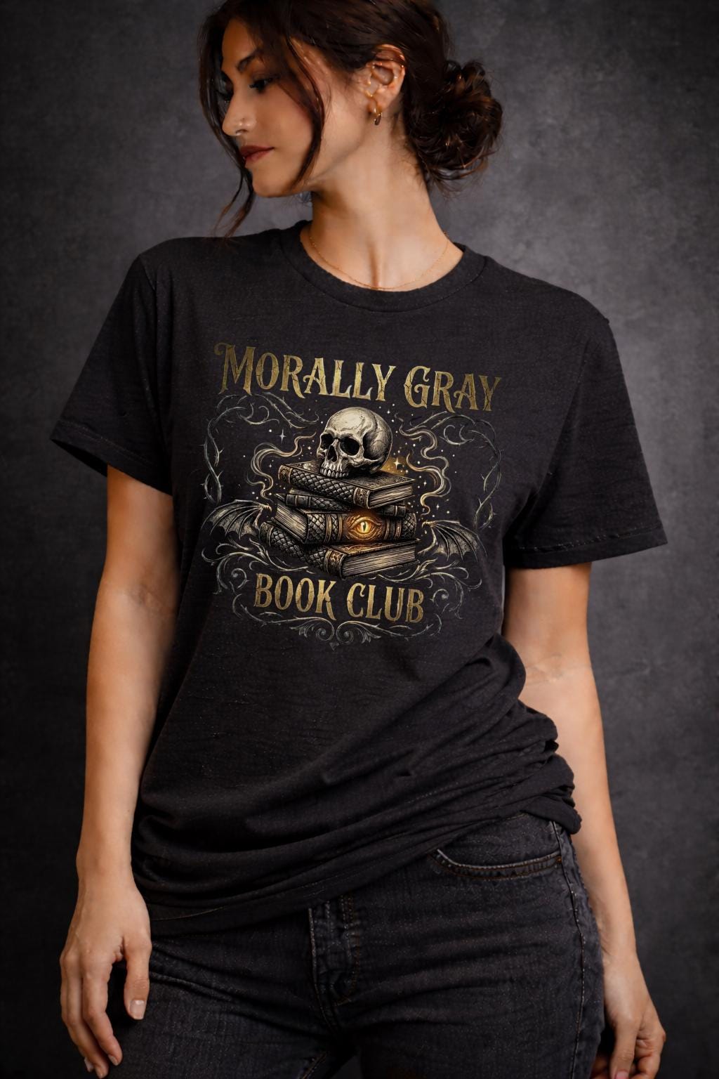 Woman wearing a black “Morally Gray Book Club” graphic t-shirt featuring a skull on stacked books with a glowing eye, styled in a dark academia aesthetic against a moody textured background.