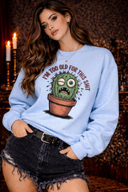 Woman wearing a light blue Gildan 18000 sweatshirt featuring a funny cactus graphic with the phrase “I’m too old for this shit,” styled casually in a cozy, warm-toned aesthetic setting.