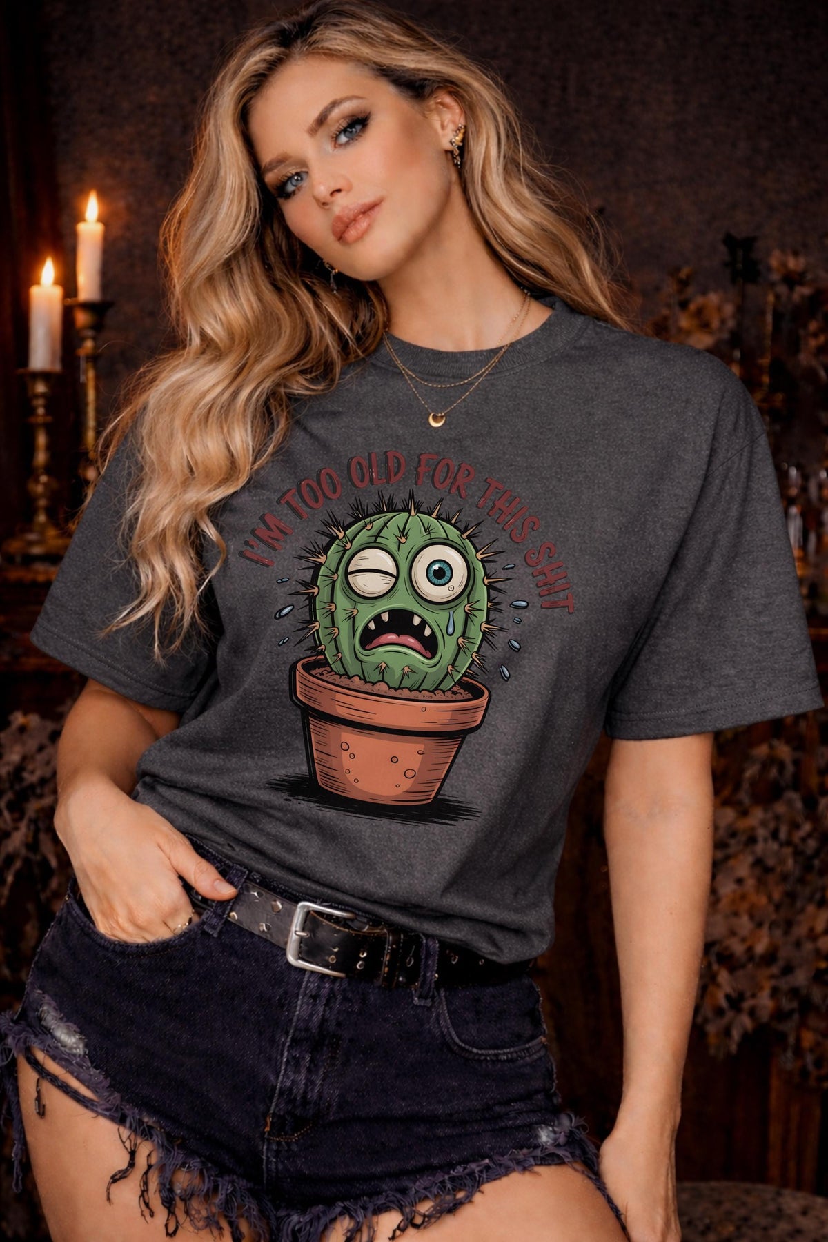 Woman wearing a Gildan 5000 t-shirt featuring a funny cactus graphic with the phrase “I’m too old for this shit,” styled with black distressed shorts in a warm, dark aesthetic setting.