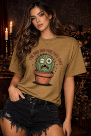 Woman wearing a Old Gold Gildan 5000 t-shirt featuring a funny cactus graphic with the phrase “I’m too old for this shit,” styled with black distressed shorts in a warm, dark aesthetic setting.