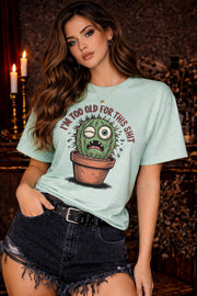 Woman wearing a Mint Gildan 5000 t-shirt featuring a funny cactus graphic with the phrase “I’m too old for this shit,” styled with black distressed shorts in a warm, dark aesthetic setting.