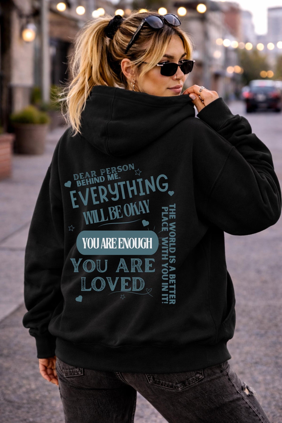 Woman wearing a black hoodie with a positive mental health message on the back reading “Dear person behind me everything will be okay you are enough you are loved” in a casual street setting.