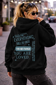 Woman wearing a black hoodie with a positive mental health message on the back reading “Dear person behind me everything will be okay you are enough you are loved” in a casual street setting.