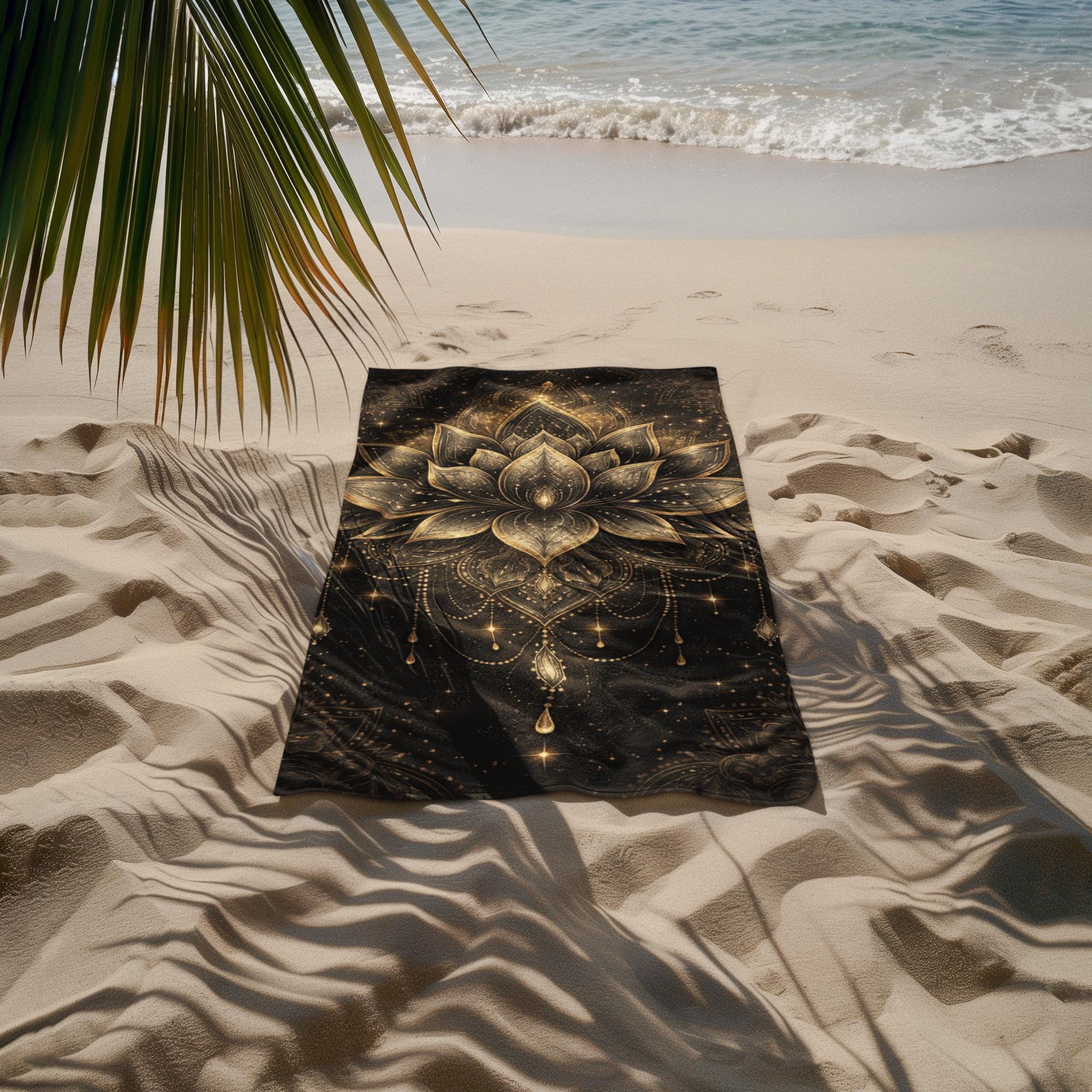 Black and gold lotus beach towel with mandala design laid on sandy beach near ocean with palm shadow
