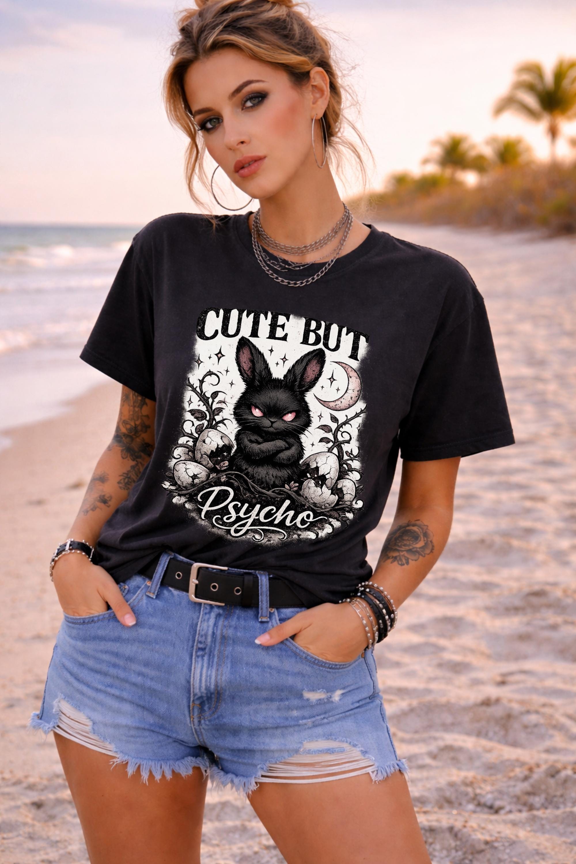 Black graphic t-shirt featuring gothic bunny with glowing eyes and “Cute But Psycho” text, worn by model on beach with casual summer outfit