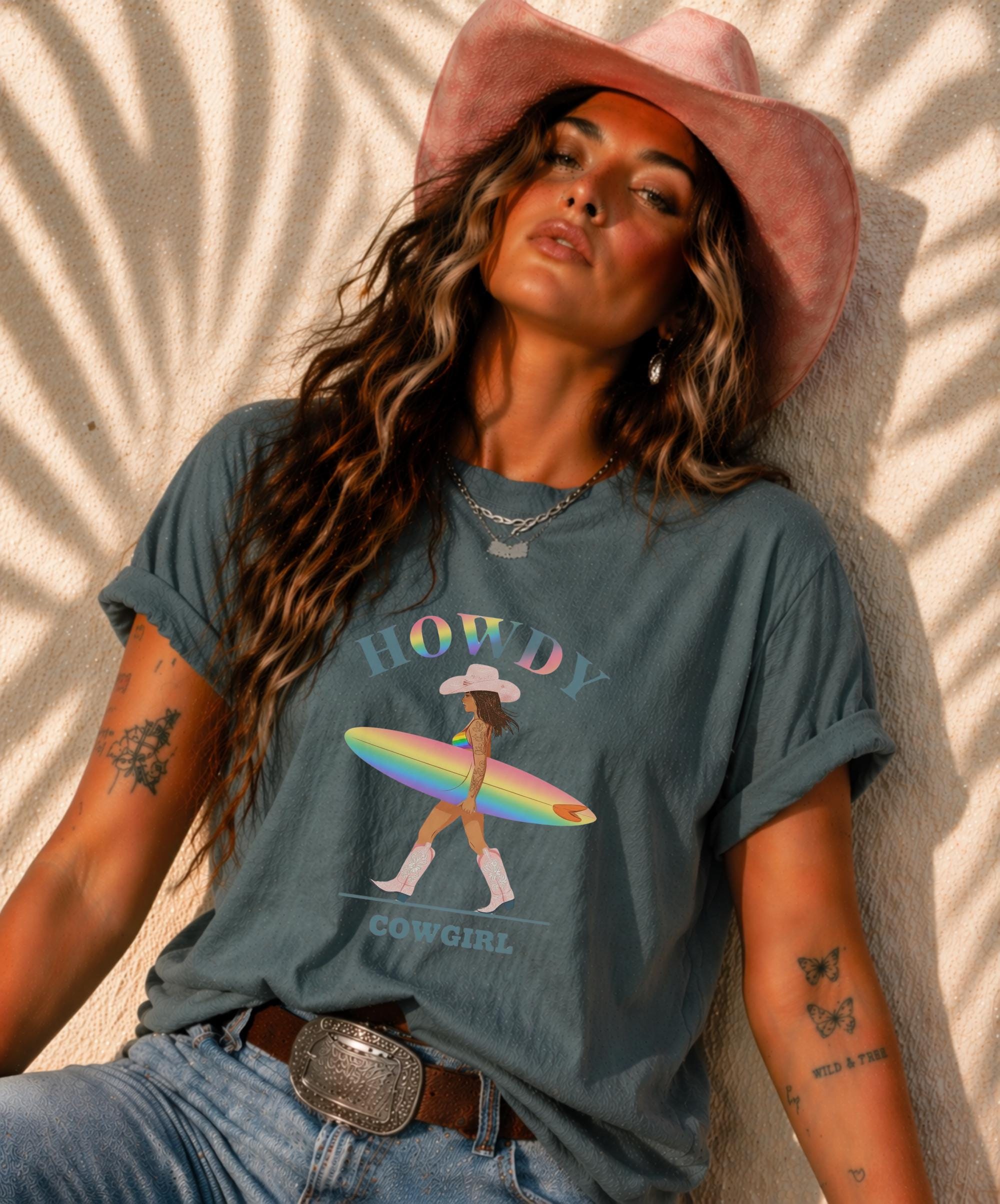 Woman wearing Comfort Colors Tshirt with Howdy Cowgirl rainbow surf design, beach sunset background, boho western outfit