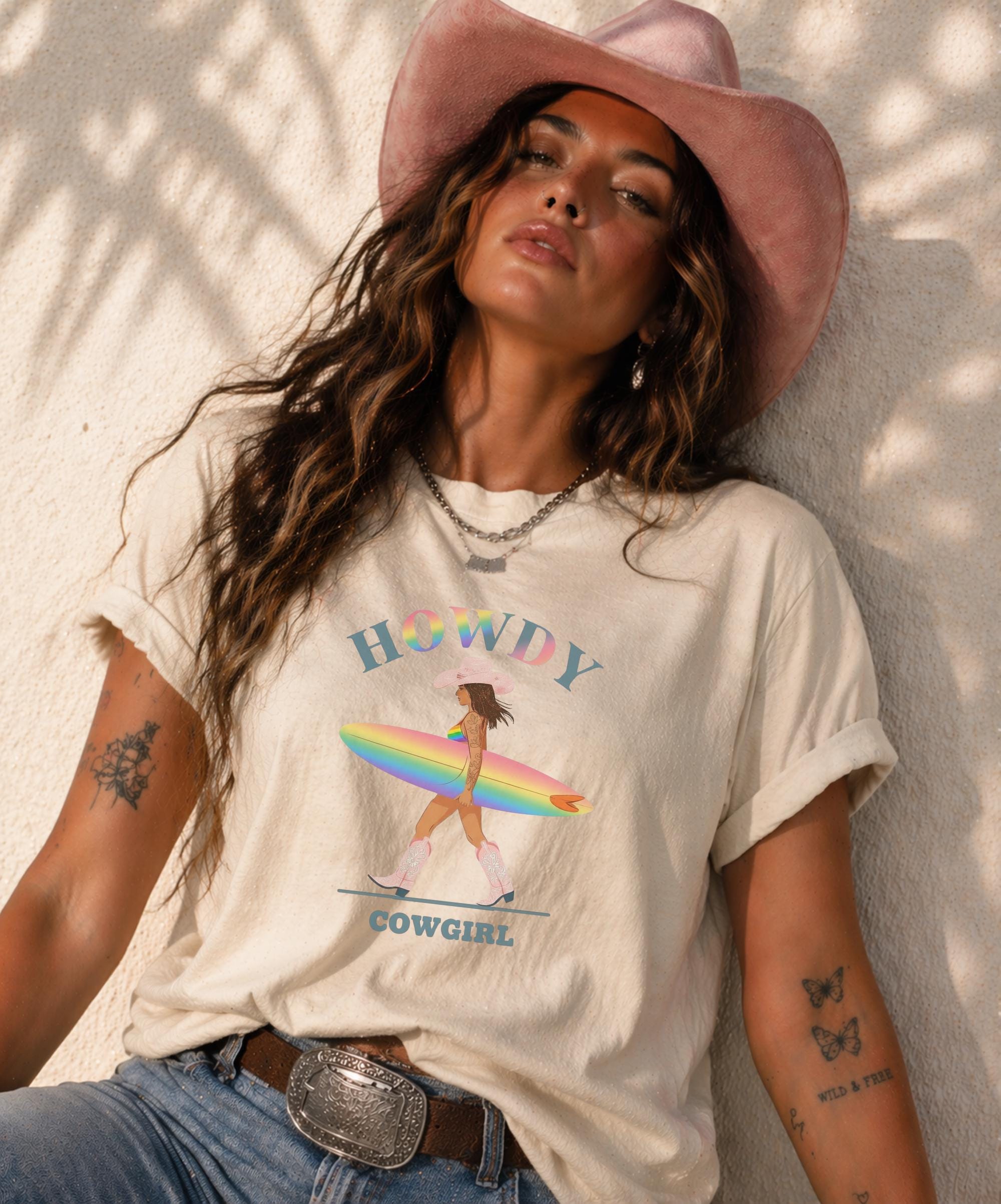 Woman wearing Comfort Colors Tshirt with Howdy Cowgirl rainbow surf design, beach sunset background, boho western outfit