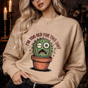 Woman wearing a sand Gildan 18000 sweatshirt featuring a funny cactus graphic with the phrase “I’m too old for this shit,” styled casually in a cozy, warm-toned aesthetic setting.