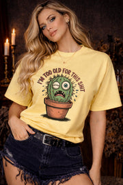 Woman wearing a Gildan 5000 t-shirt featuring a funny cactus graphic with the phrase “I’m too old for this shit,” styled with black distressed shorts in a warm, dark aesthetic setting.