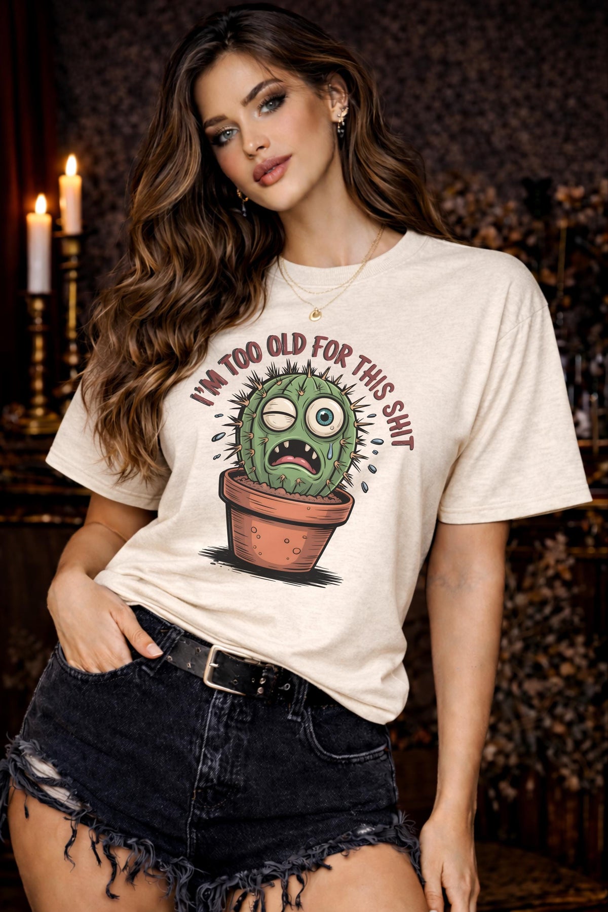 Woman wearing a Gildan 5000 t-shirt featuring a funny cactus graphic with the phrase “I’m too old for this shit,” styled with black distressed shorts in a warm, dark aesthetic setting.