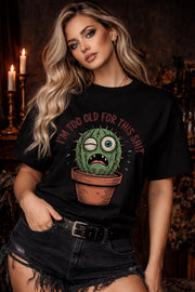 Woman wearing a black Gildan 5000 t-shirt featuring a funny cactus graphic with the phrase “I’m too old for this shit,” styled with black distressed shorts in a warm, dark aesthetic setting.