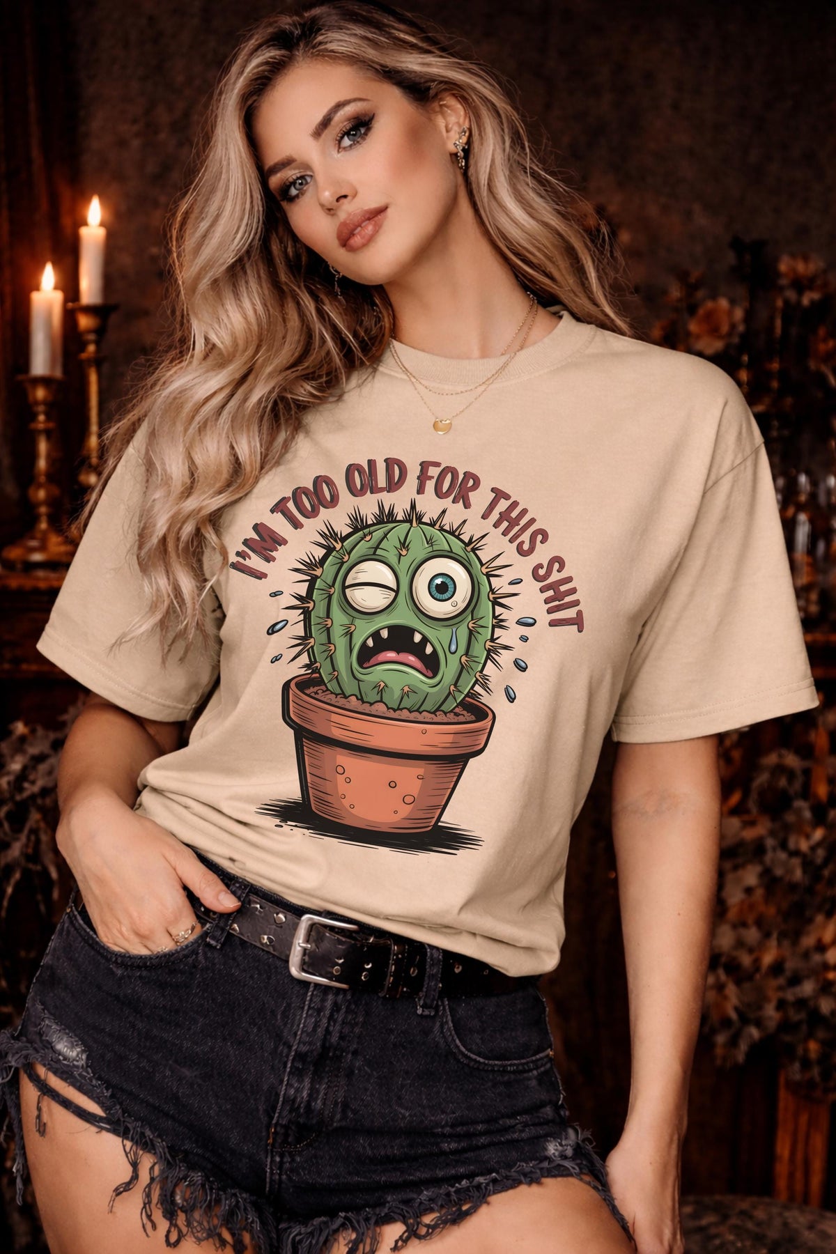Woman wearing a beige Gildan 5000 t-shirt featuring a funny cactus graphic with the phrase “I’m too old for this shit,” styled with black distressed shorts in a warm, dark aesthetic setting.