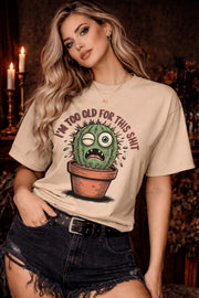 Woman wearing a beige Gildan 5000 t-shirt featuring a funny cactus graphic with the phrase “I’m too old for this shit,” styled with black distressed shorts in a warm, dark aesthetic setting.