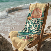 Stay salty beach towel with seashell and ocean wave design draped over chair on sandy beach by ocean