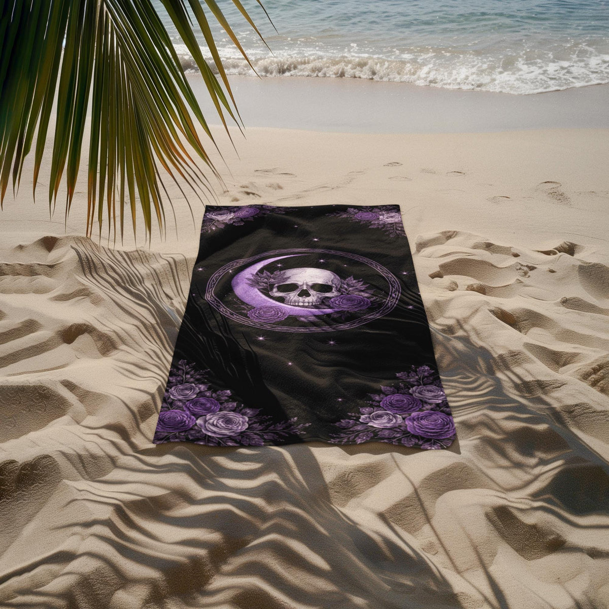 Purple gothic skull beach towel with moon and floral design laid on sandy beach near ocean with palm shadow