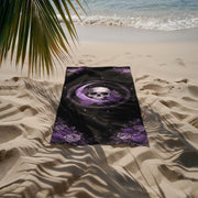 Purple gothic skull beach towel with moon and floral design laid on sandy beach near ocean with palm shadow