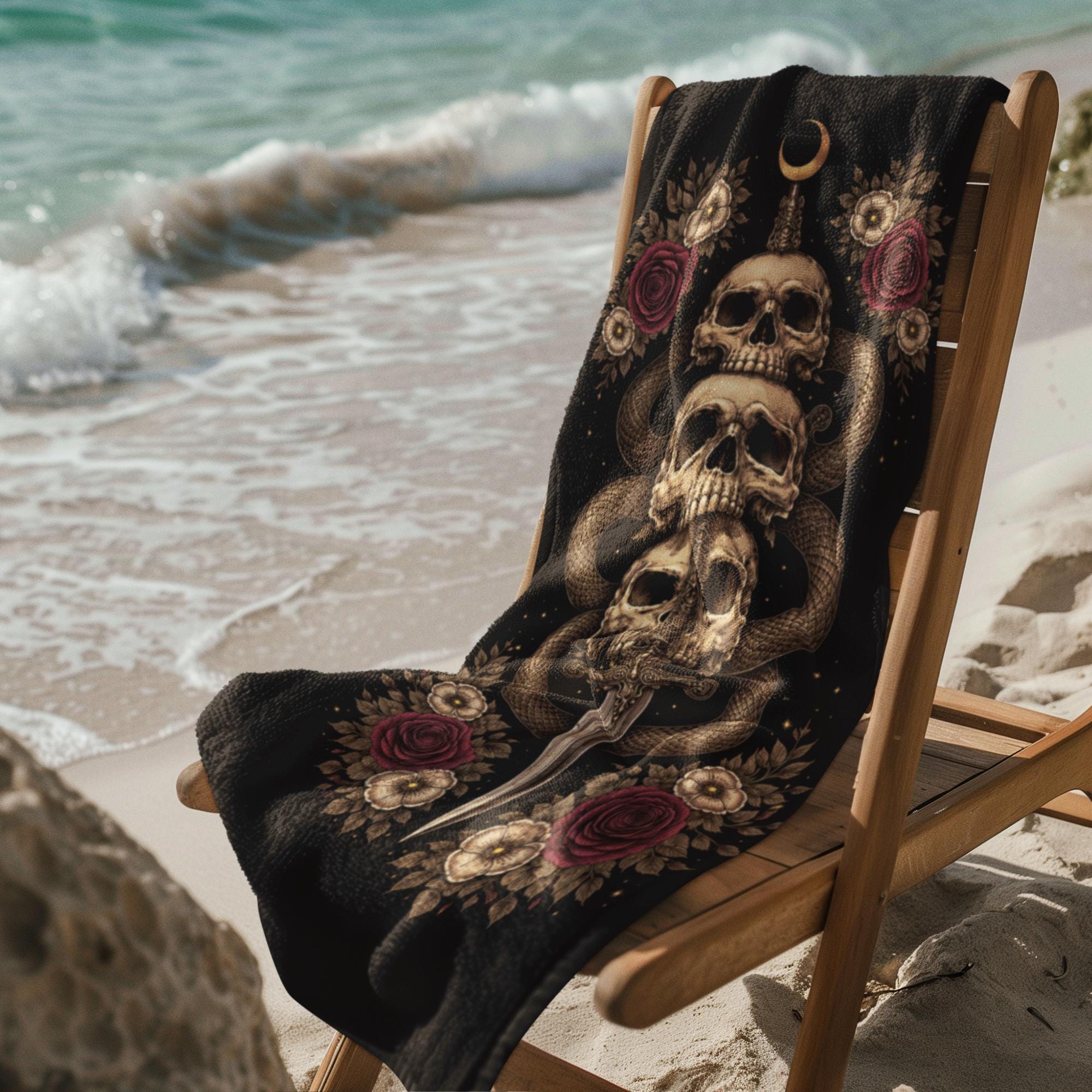 Gothic skull beach towel with snake dagger and floral design draped over chair on sandy beach by ocean