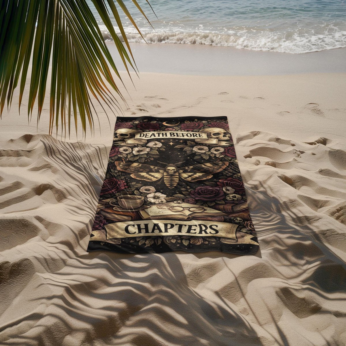 Dark gothic beach towel with death before chapters design featuring skulls moth and books laid on sandy beach near ocean