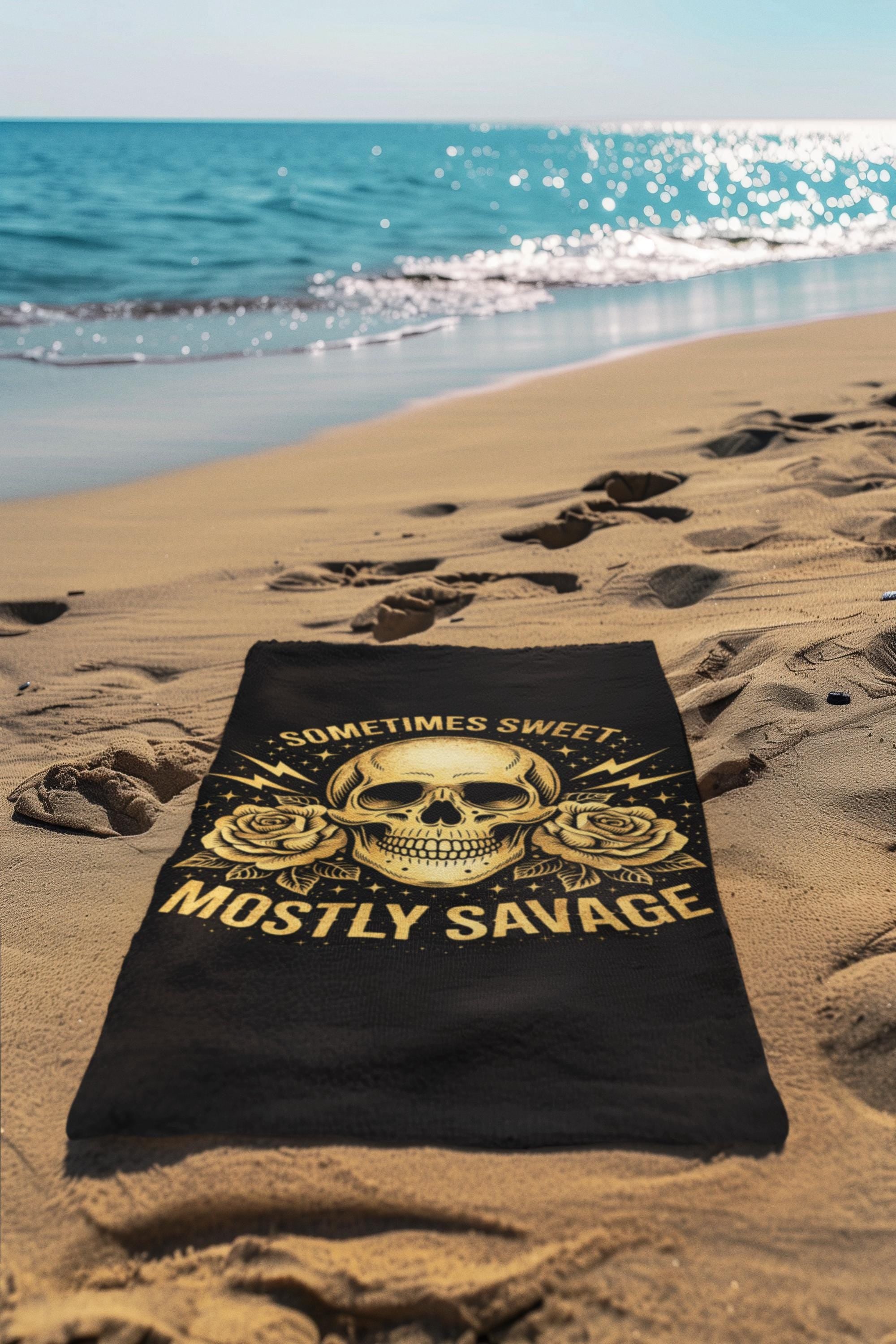 Black beach towel with skull design and text sometimes sweet mostly savage laid on sandy beach near ocean