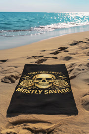 Black beach towel with skull design and text sometimes sweet mostly savage laid on sandy beach near ocean