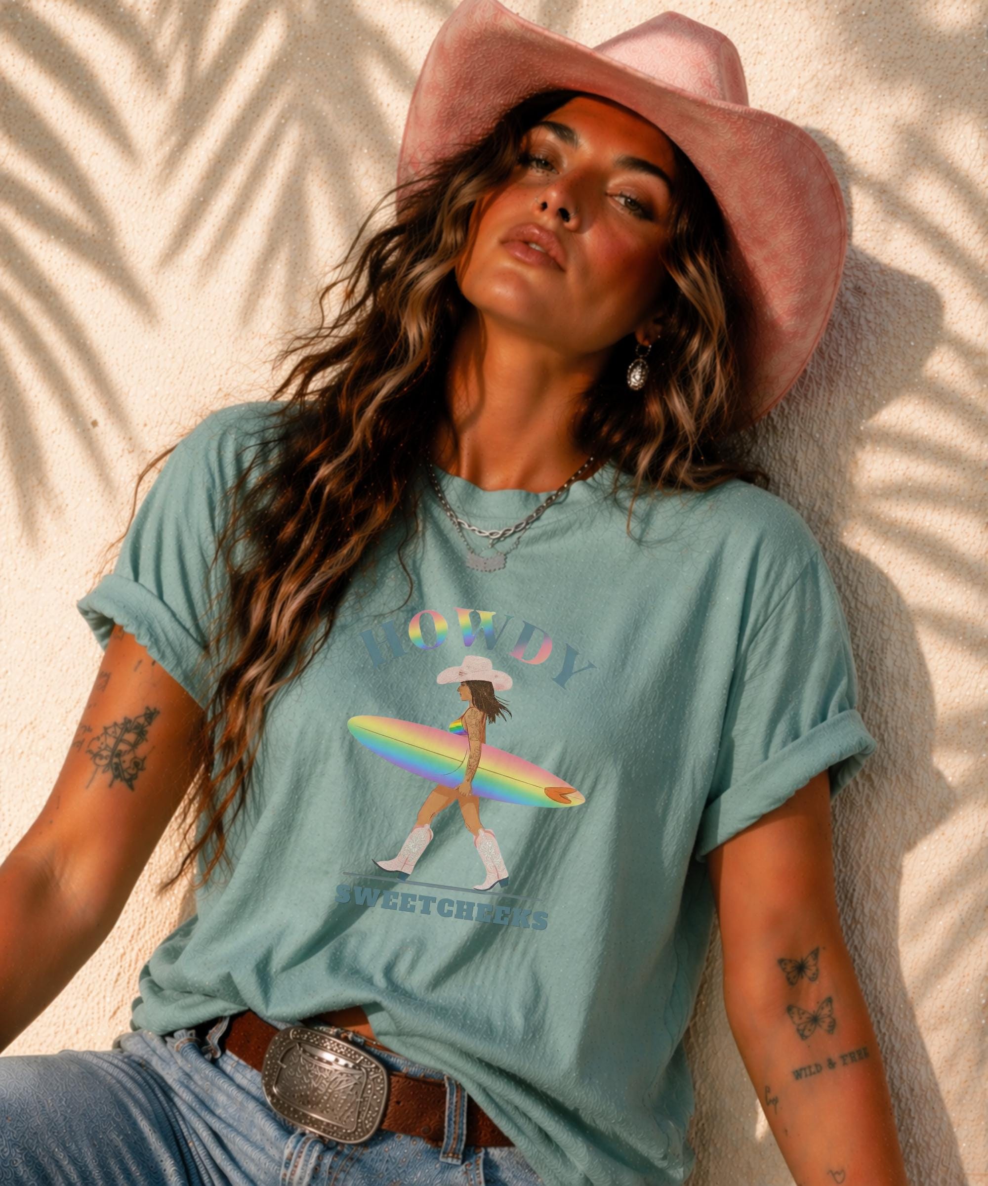 Woman wearing Comfort Colors Tshirt with Howdy Sweetcheeks rainbow surf design, beach sunset background, boho western outfit