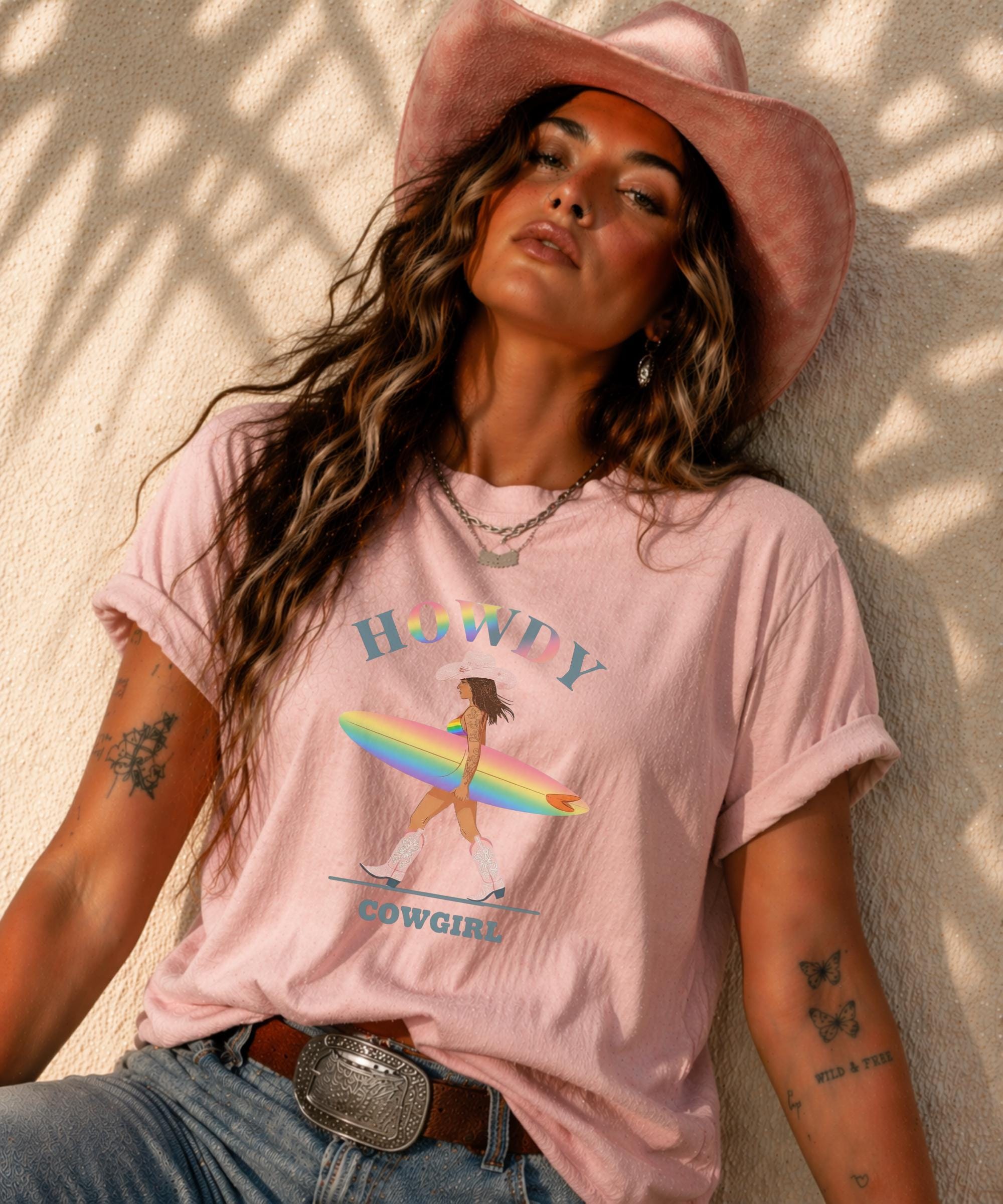 Woman wearing Comfort Colors Tshirt with Howdy Cowgirl rainbow surf design, beach sunset background, boho western outfit