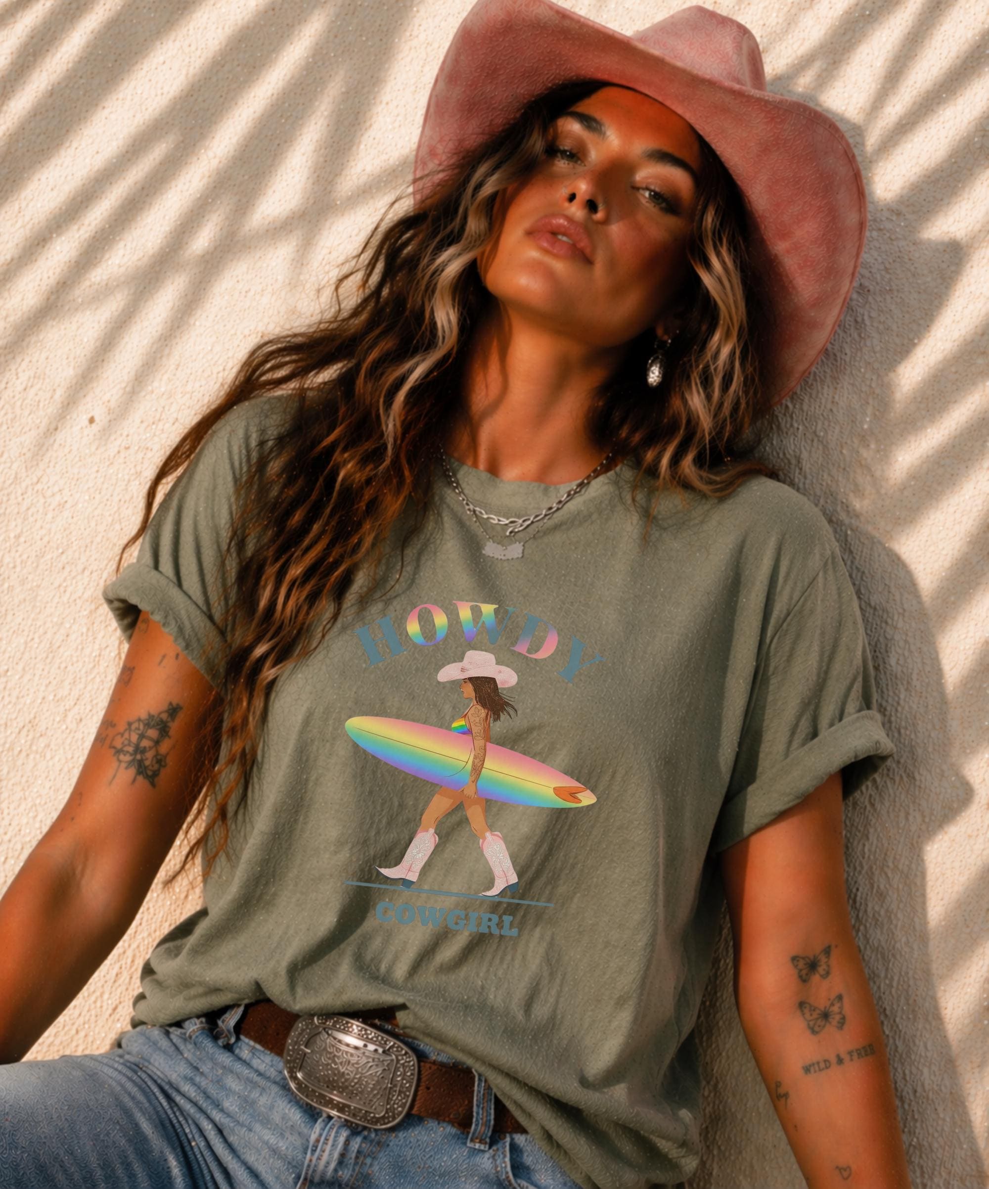 Woman wearing Comfort Colors Tshirt with Howdy Cowgirl rainbow surf design, beach sunset background, boho western outfit
