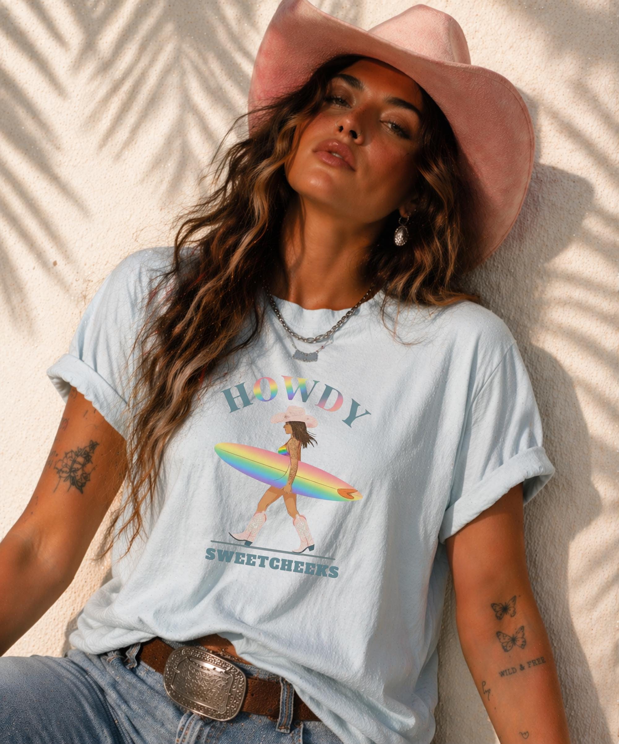 Woman wearing Comfort Colors Tshirt with Howdy SweetCheeks rainbow surf design, beach sunset background, boho western outfit
