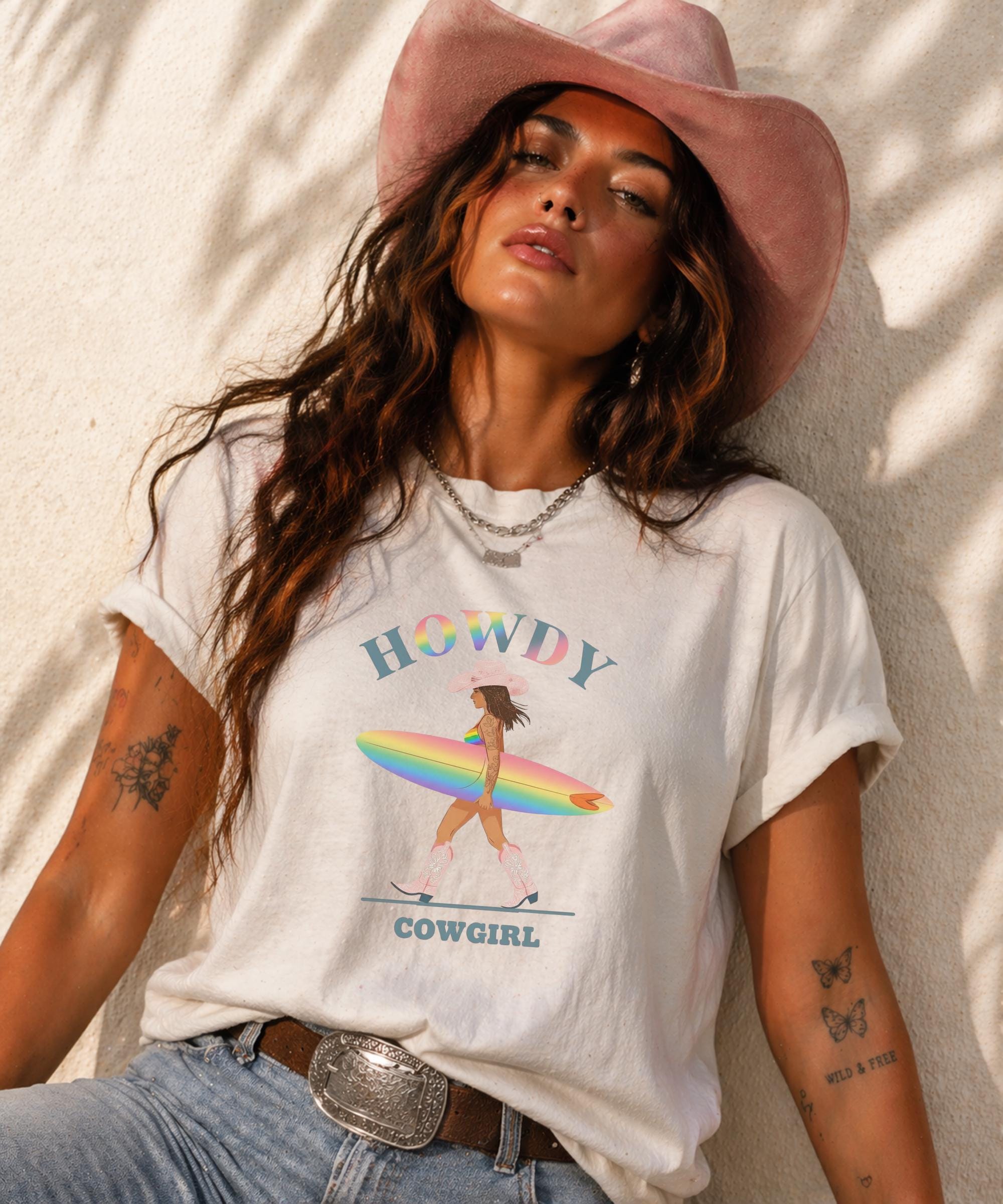 Woman wearing Comfort Colors Tshirt with Howdy Cowgirl rainbow surf design, beach sunset background, boho western outfit