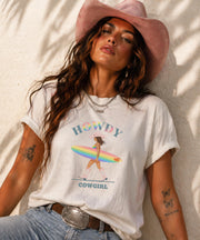 Woman wearing Comfort Colors Tshirt with Howdy Cowgirl rainbow surf design, beach sunset background, boho western outfit