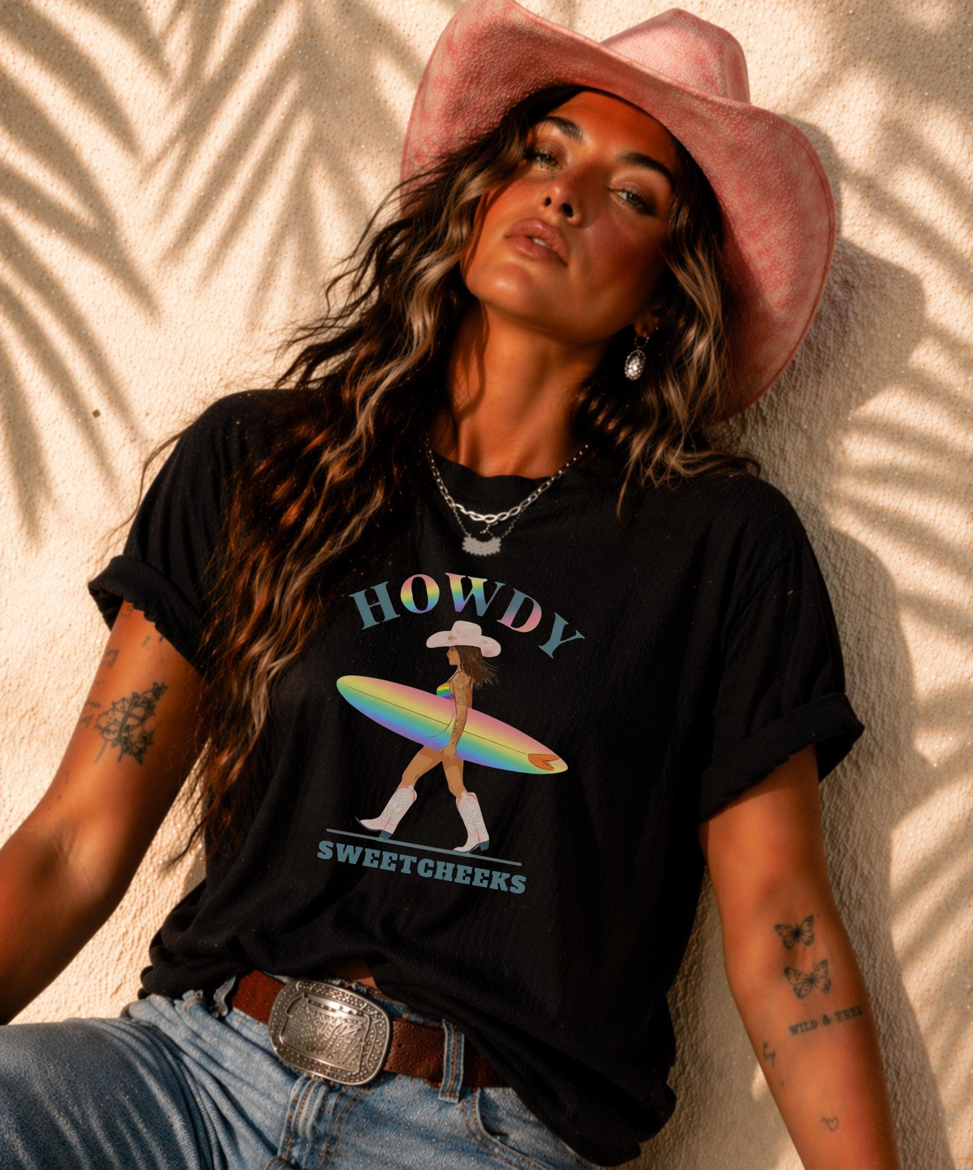 Woman wearing Comfort Colors tank top with Howdy Sweetcheeks rainbow surf design, beach sunset background, boho western outfit