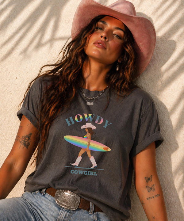 Woman wearing Comfort Colors Tshirt with Howdy Cowgirl rainbow surf design, beach sunset background, boho western outfit