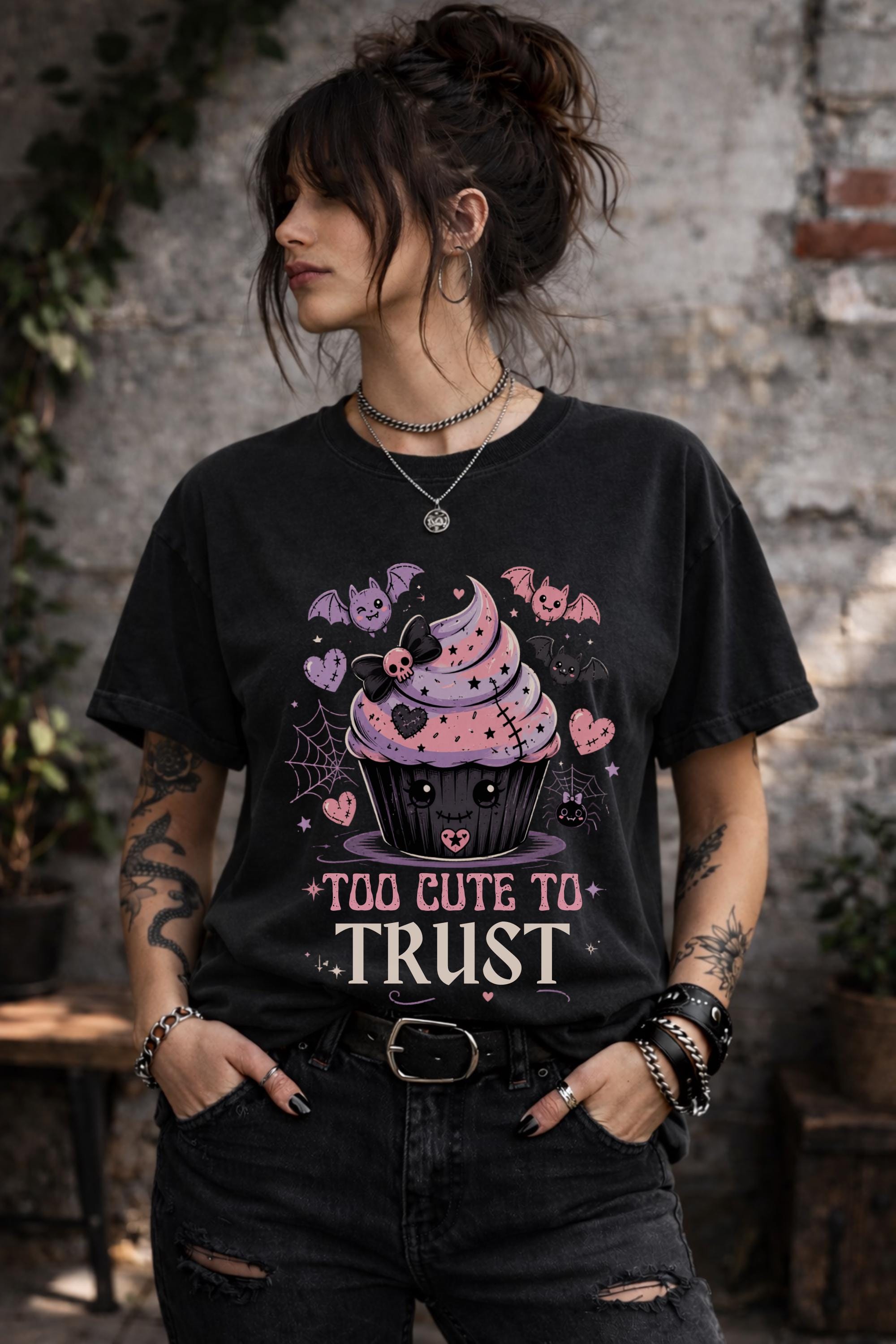 Woman wearing a graphic t-shirt featuring spooky kawaii cupcake with bats hearts spiderwebs and Too Cute to Trust quote