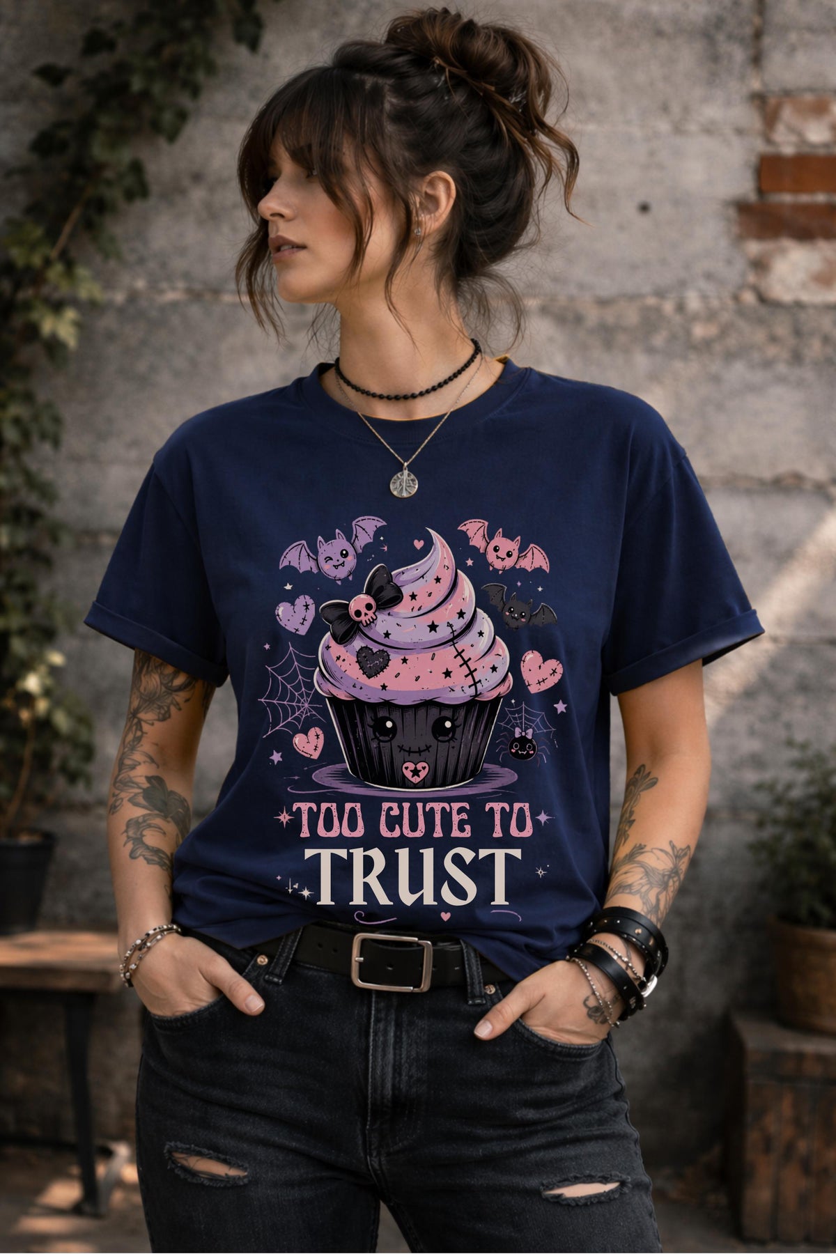 Woman wearing a graphic t-shirt featuring spooky kawaii cupcake with bats hearts spiderwebs and Too Cute to Trust quote