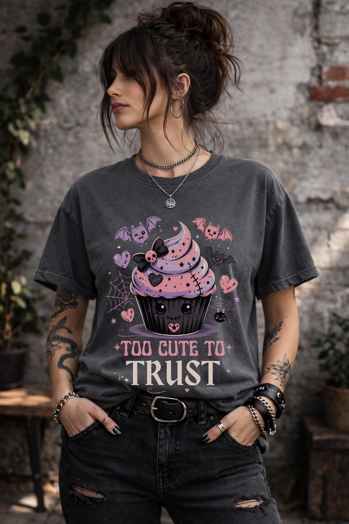 Woman wearing a graphic t-shirt featuring spooky kawaii cupcake with bats hearts spiderwebs and Too Cute to Trust quote