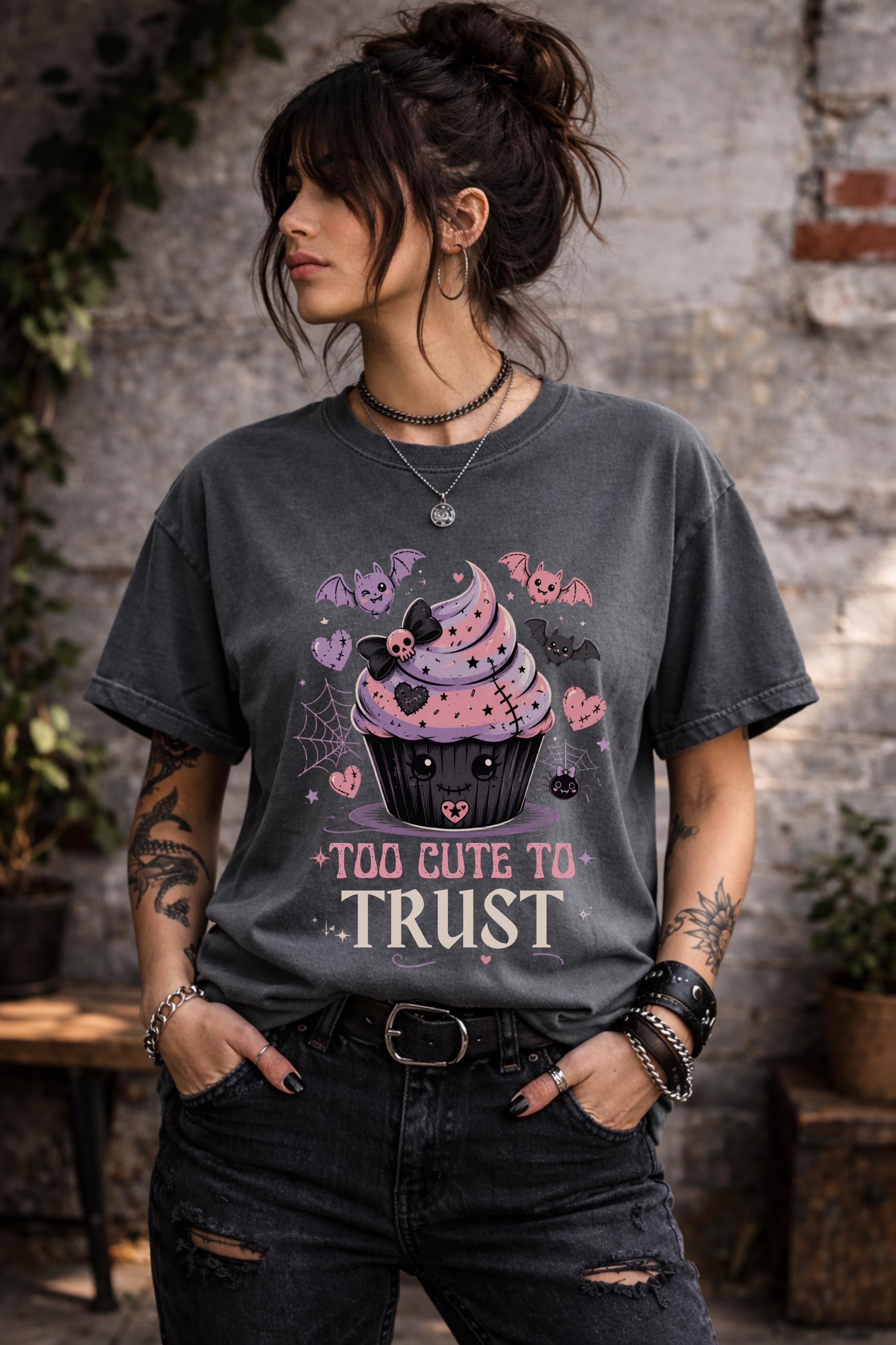 Woman wearing a graphic t-shirt featuring spooky kawaii cupcake with bats hearts spiderwebs and Too Cute to Trust quote
