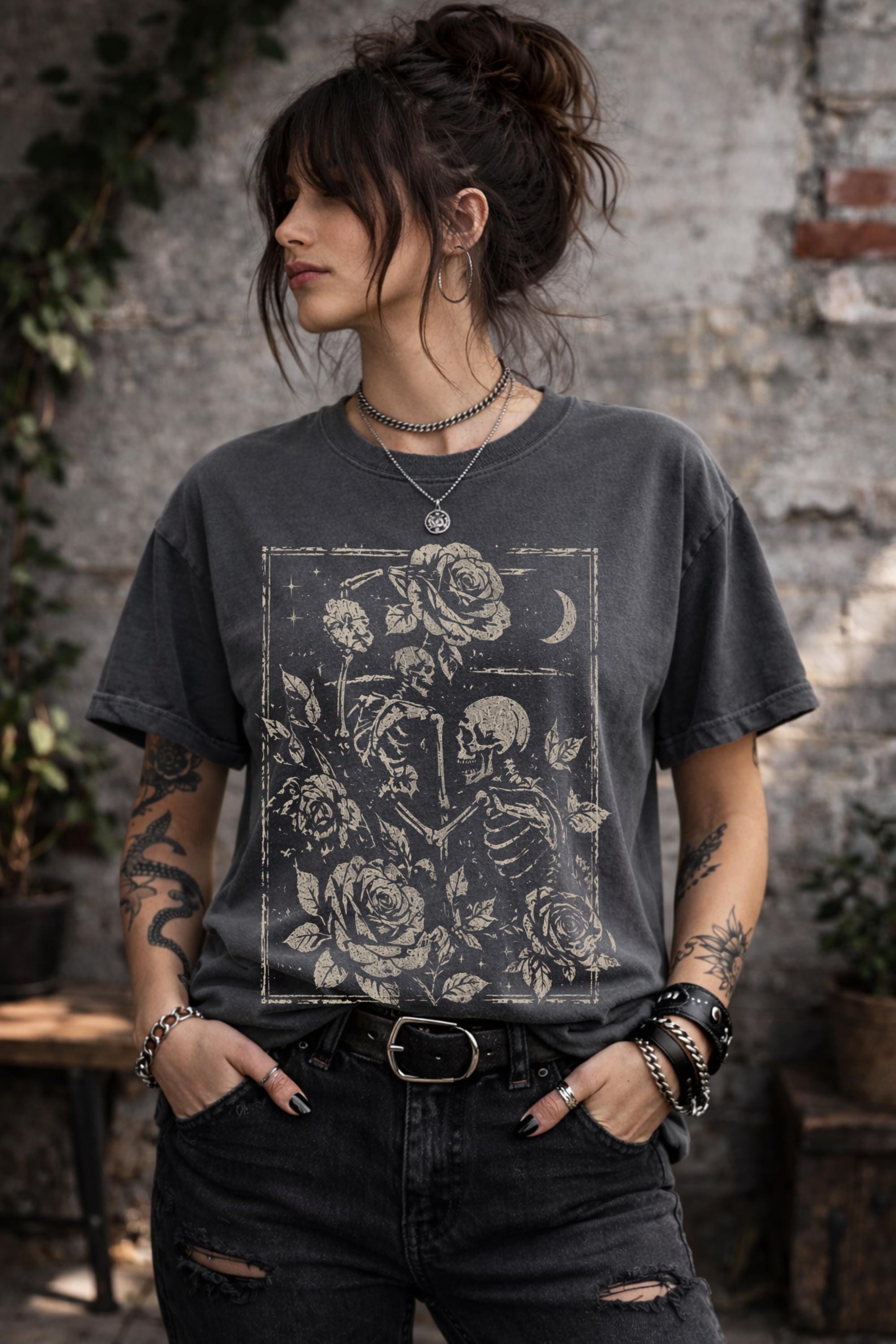 Woman wearing a graphic t-shirt featuring two skeleton lovers surrounded by roses beneath a crescent moon in a gothic dark academia style design.