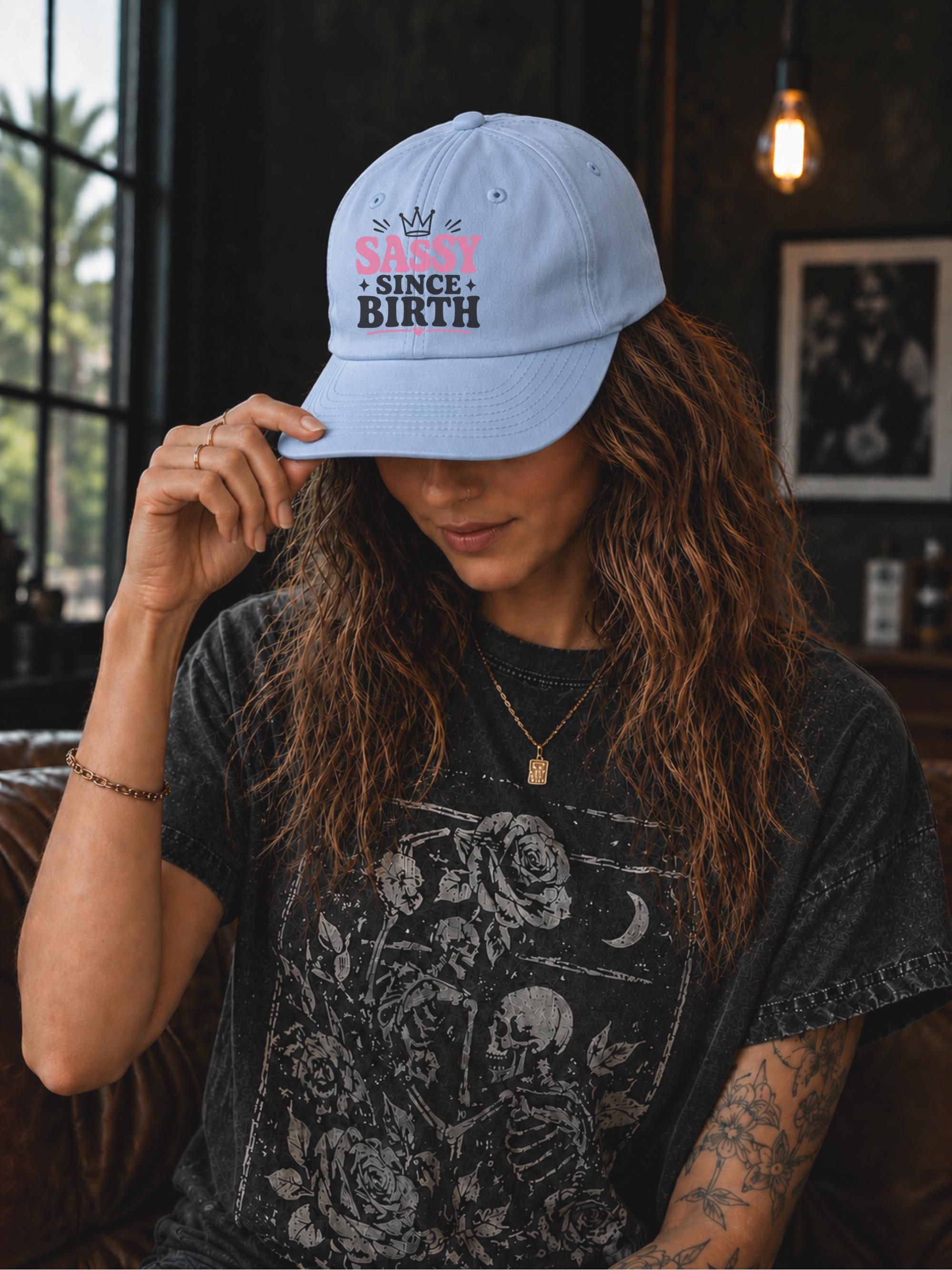 Classic dad cap featuring a pink and white "Sassy Since Birth" graphic with crown artwork, styled in a casual fashion setting.