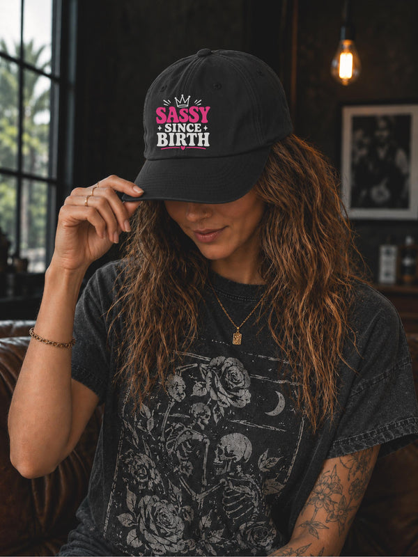 Classic dad cap featuring a pink and white "Sassy Since Birth" graphic with crown artwork, styled in a casual fashion setting.